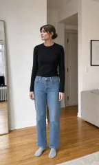 A woman standing in her apartment in a black top and blue jeans
