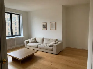 A bright apartment living room with a cream sofa, small coffee table, two undersized prints, and an empty corner